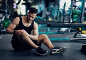 Sport Trauma. Young Arab Male Athlete Touching His Hurted Knee At Gym, Upset Middle Eastern Guy Sitting On Floor And Massaging Sore Injured Leg After Workout Training In Fitness Club, Free Space
